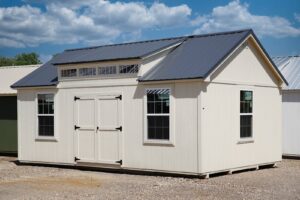 A white shed with a metal roof sitting on a gravel lot available for sale.
