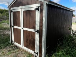 A brown and white 8x12 Utility Shed for sale in a field.