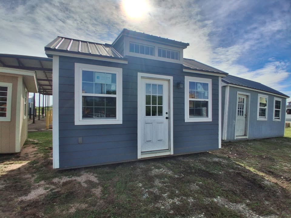  - Front view of a blue modular house with white-trimmed windows and a central white door under a sunny sky.