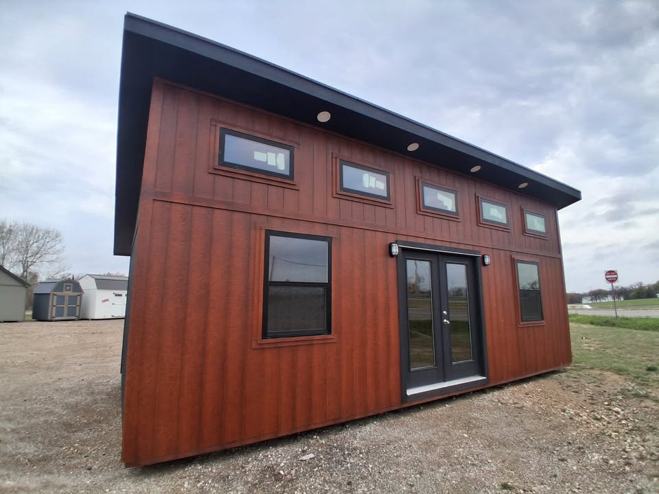  - Rust-colored metal tiny house with a black-framed double door, several small upper windows, and a slanted roof on a gravel lot under an overcast sky.