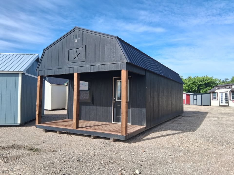  - Small dark gray wooden building with a covered front porch on a wooden deck, supported by two brown posts, in a gravel lot under a blue sky.
