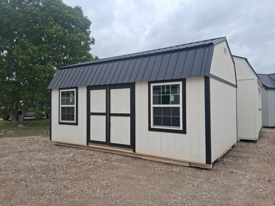 - Small beige storage shed with dark trim and a metal gabled roof on a gravel lot, featuring double doors and two windows with black frames.