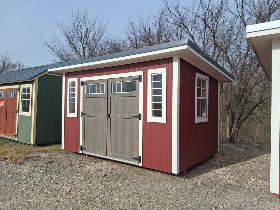  - Red shed with white trim and double gray doors in a row of colorful storage sheds on a gravel lot, bare trees in background