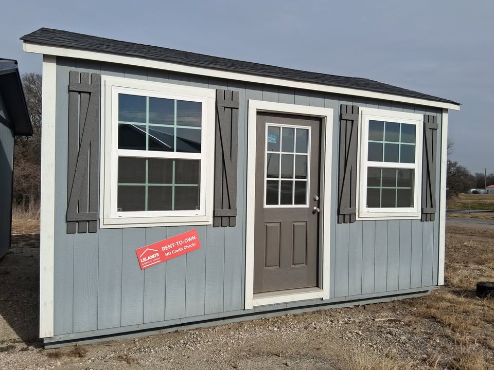  - Small gray modular home with two white-framed windows, dark shutters, and a brown front door on a gravel lot.