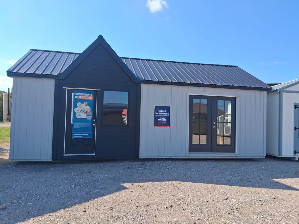  - Modular building on a gravel lot with a peaked front entrance, dark doorway, and two large glass doors; posters are visible on the exterior walls.