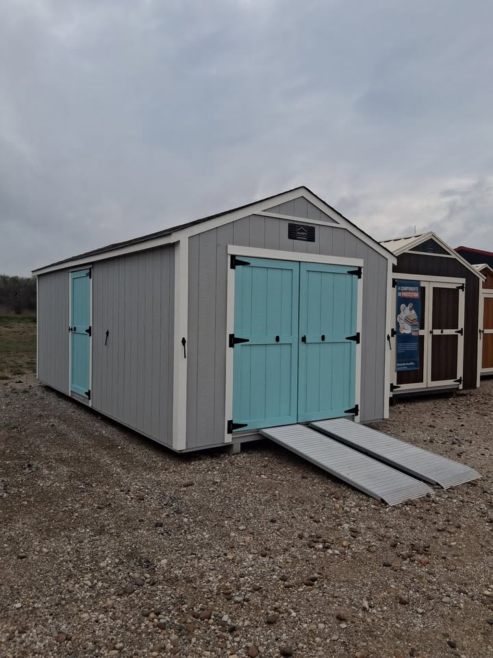  - Gray storage shed with light blue double doors and a metal ramp on a rocky lot, under cloudy sky.