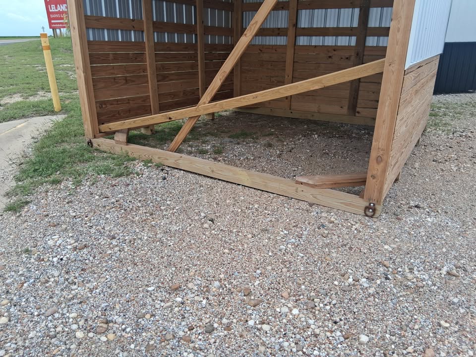  - Partially assembled wooden shed frame on a gravel lot; the bottom beam is cracked and detached at a corner with diagonal braces visible.
