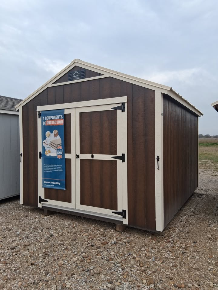  - Brown storage shed with white trim and double doors on a gravel lot; promotional sign on the left door.