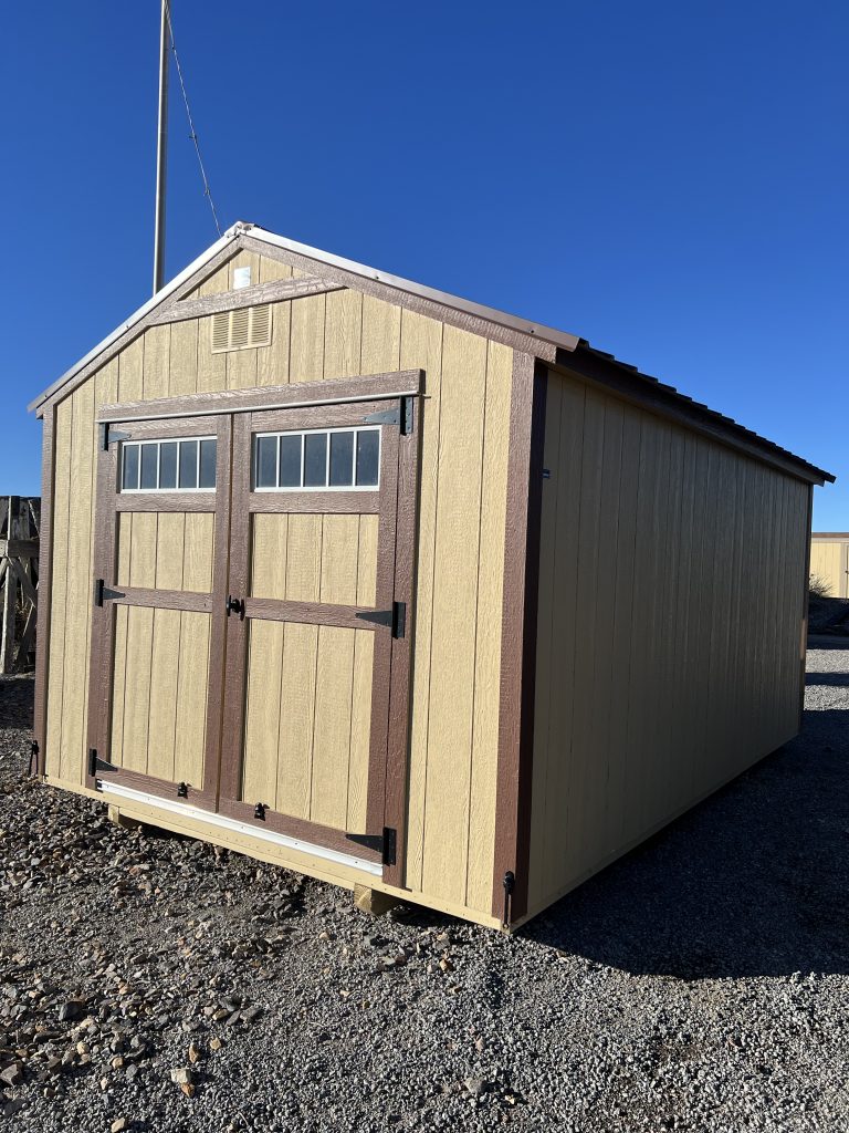  - Beige wooden storage shed with brown trim and double barn-style doors on a gravel lot under a clear blue sky, featuring small window panes in the doors.