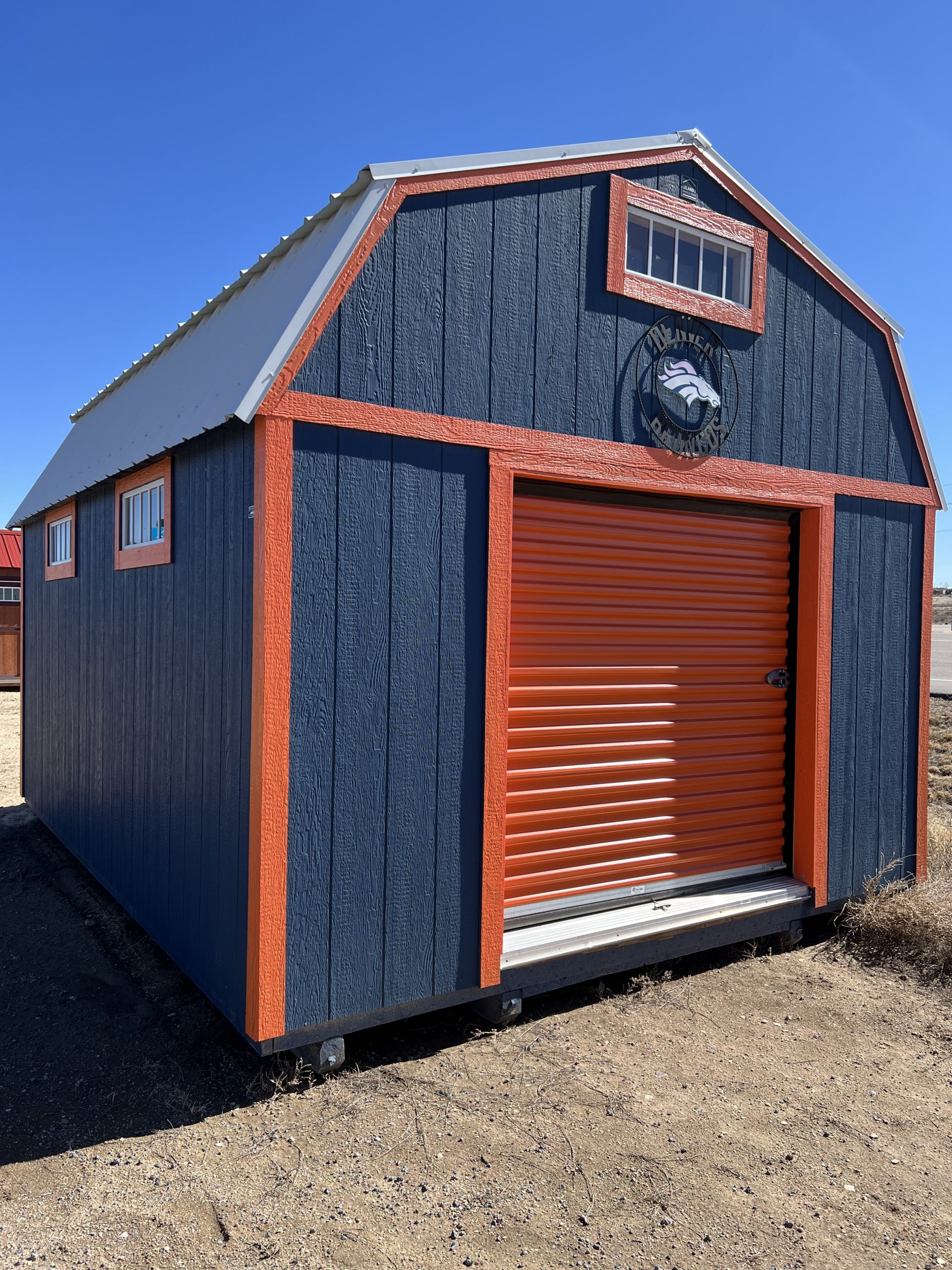  - Blue wooden storage shed with orange trim and a bright orange roll-up door, set on dry, sandy ground under a clear blue sky.