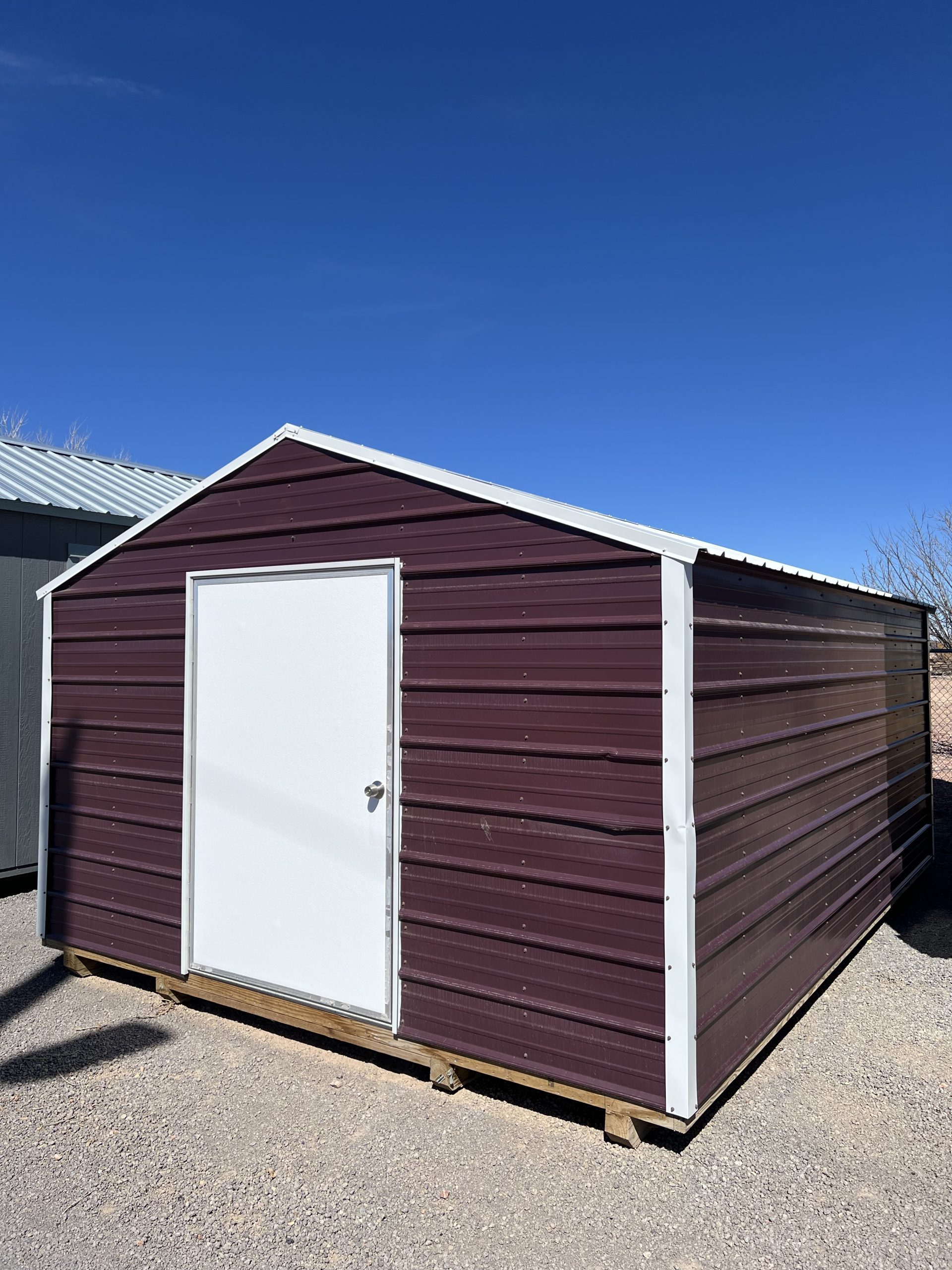  - Maroon metal storage shed with a white door, on a wooden pallet, under a clear blue sky on gravel ground.