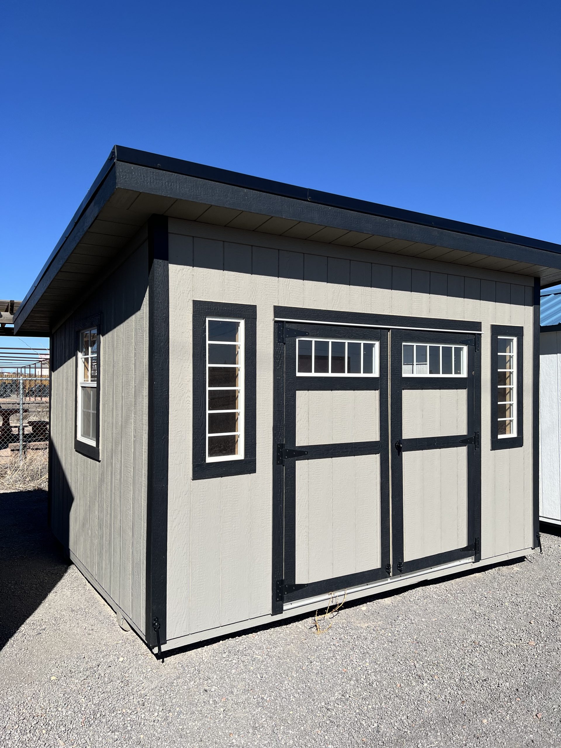  - Gray wooden storage shed with black trim and double doors featuring small window panes, set on gravel under a clear blue sky.