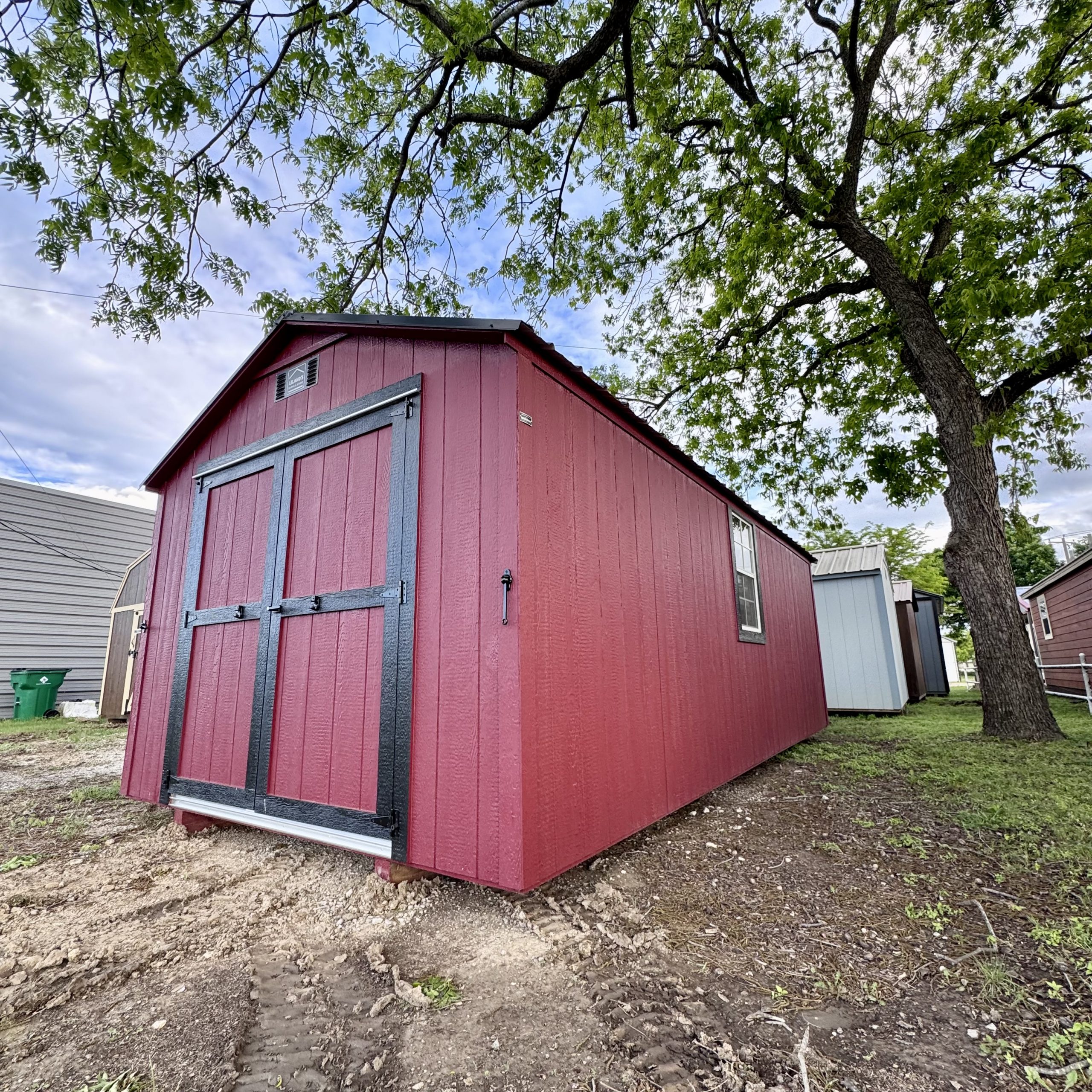  - Red wooden shed with blue trim and double doors, in a yard beneath leafy tree branches.