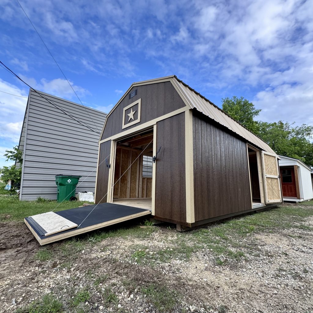  - Under-construction shed with dark brown exterior, beige trim, open doorway, and a ramp extending outward against a cloudy blue sky.