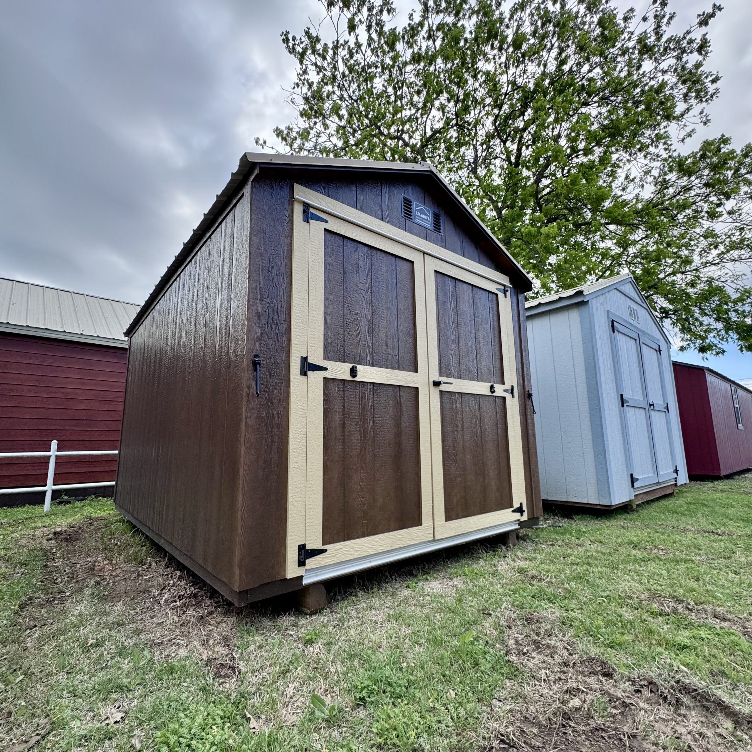  - Row of small wooden storage sheds in brown and beige, with metal roofs, set on grassy ground under a cloudy sky.