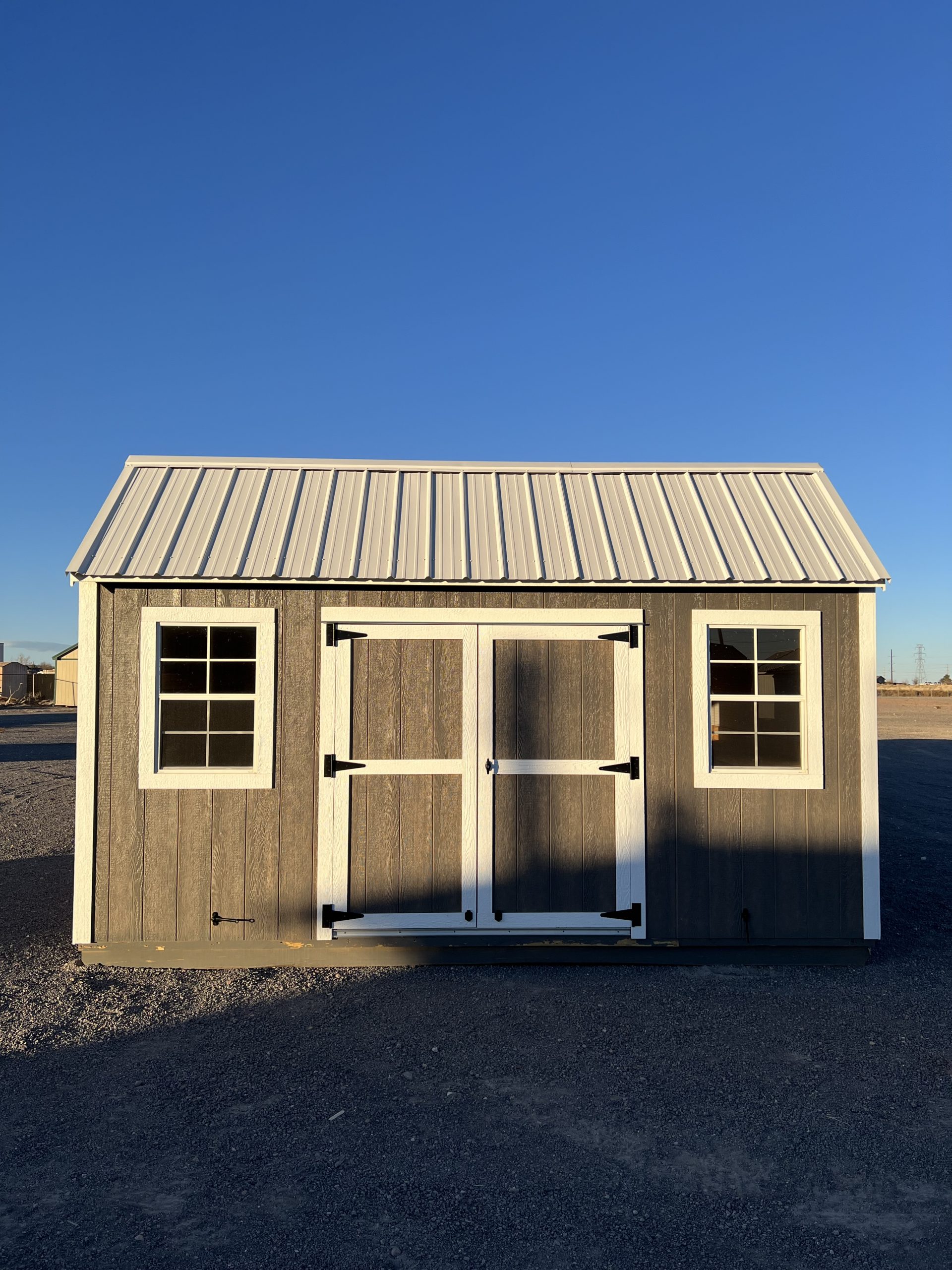  - Front view of a gray storage shed with white trim, metal roof, and two white-framed windows on a gravel lot under a clear blue sky, with double doors in the center.