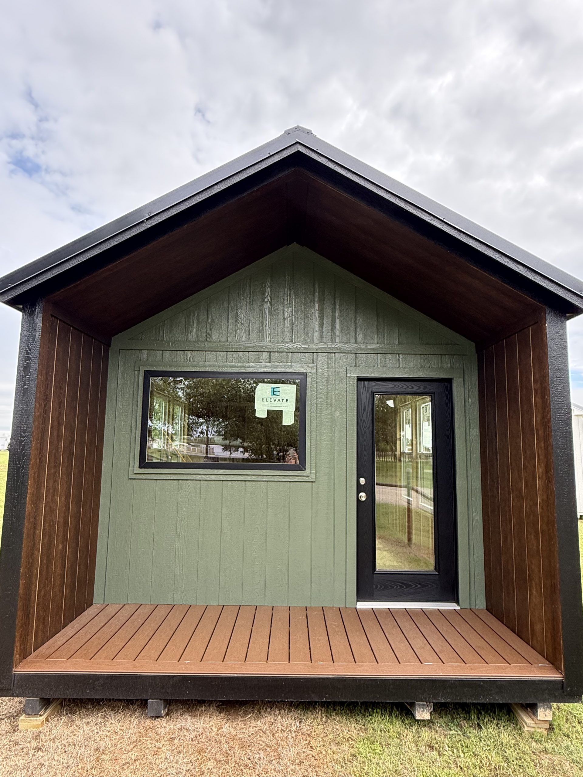 - Front view of a small green shed with a brown wooden deck and a black-framed door under a cloudy sky.