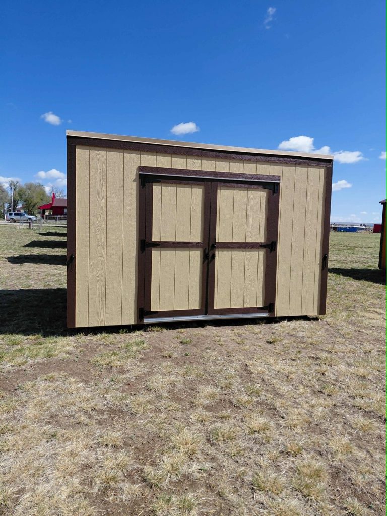  - Beige wooden storage shed with brown trim and double doors in a dry grassy field under a clear blue sky with a few clouds.