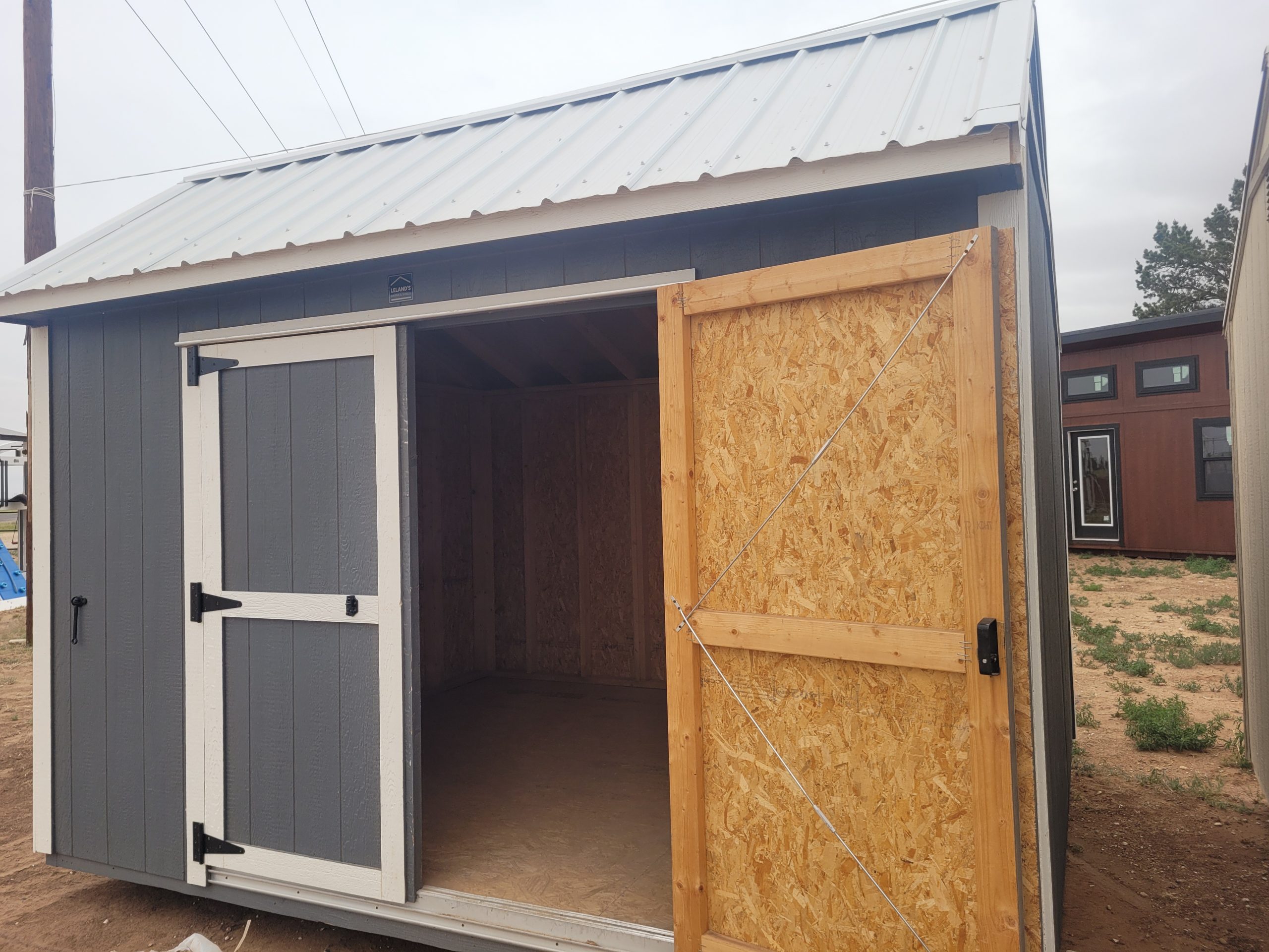 1000012355 - LELAND'S SHEDS - Small shed under construction with blue-gray siding, white trim, and a metal roof; front door open to reveal interior framing.