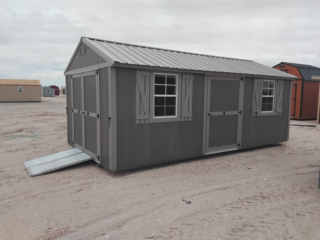 - - Gray portable storage shed with two windows and a metal ramp, on a dirt lot with other sheds in the background.