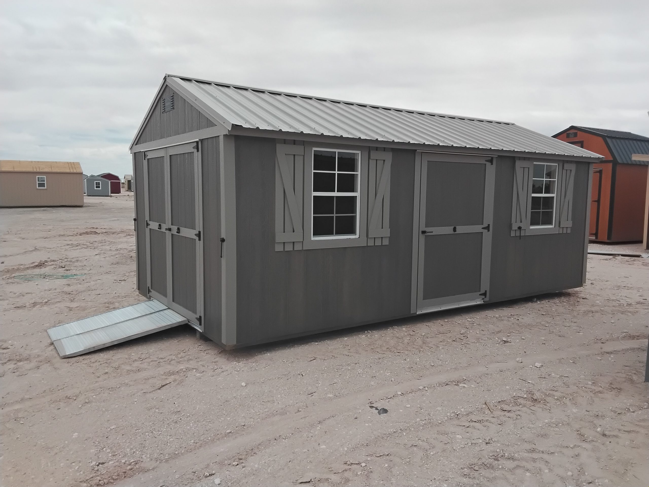 - - Gray portable storage shed with two windows and a metal ramp, on a dirt lot with other sheds in the background.