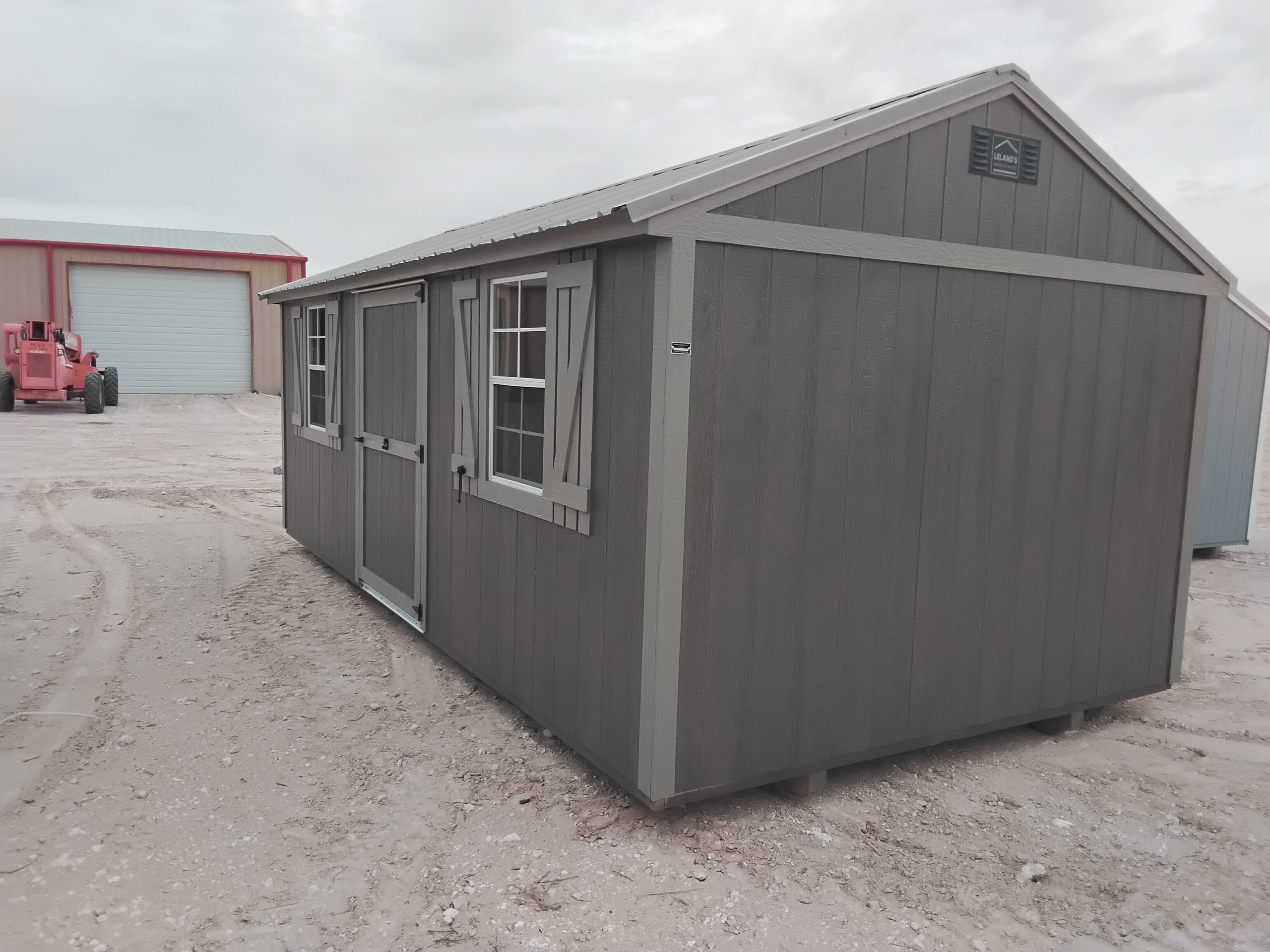 20260505_122703 - LELAND'S SHEDS - Side view of a gray wooden shed with white trim and small windows on a dirt lot, with a pink construction vehicle in the background.