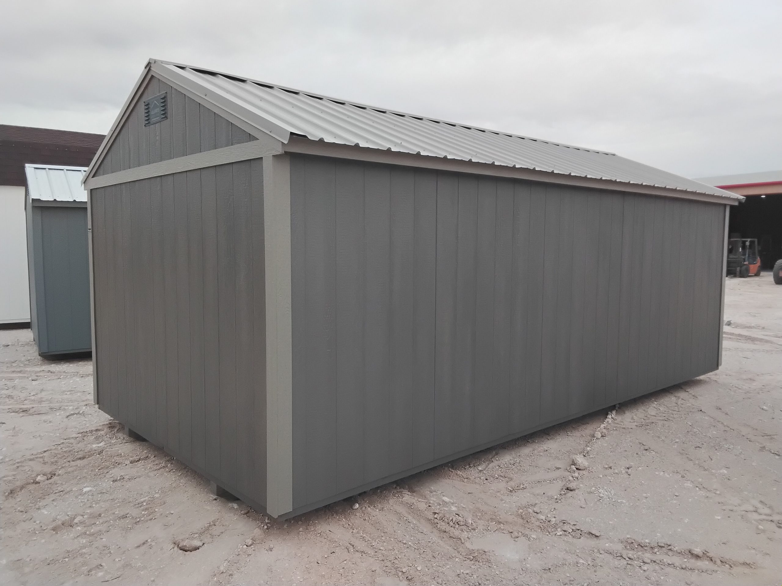 20260505_122728 - LELAND'S SHEDS - Gray corrugated metal storage shed with a pitched roof on a dirt lot; another shed is visible in the background.