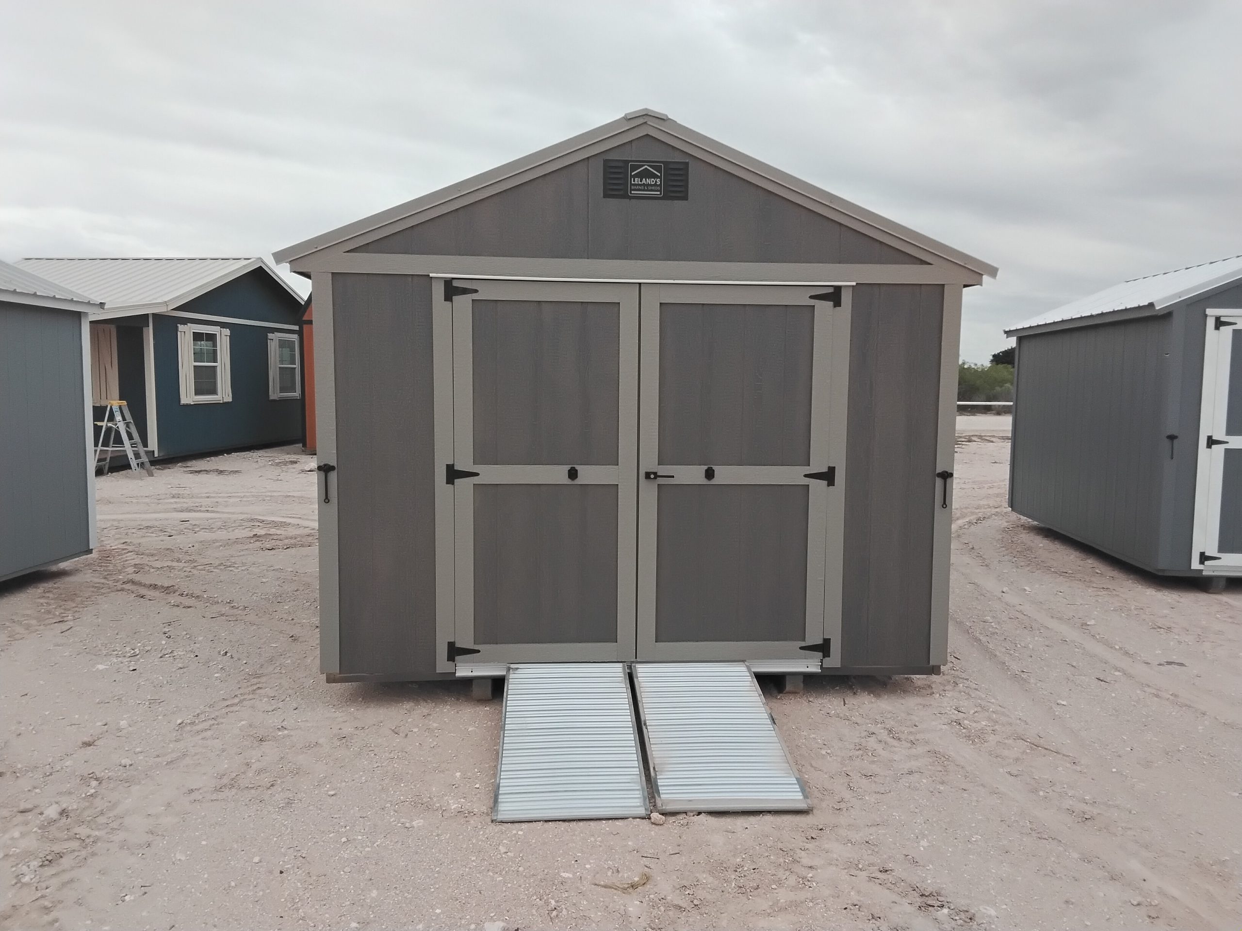 20260505_122742 - LELAND'S SHEDS - Grey storage shed with double doors on a dirt lot; metal ramp extends toward the viewer in front of the shed.
