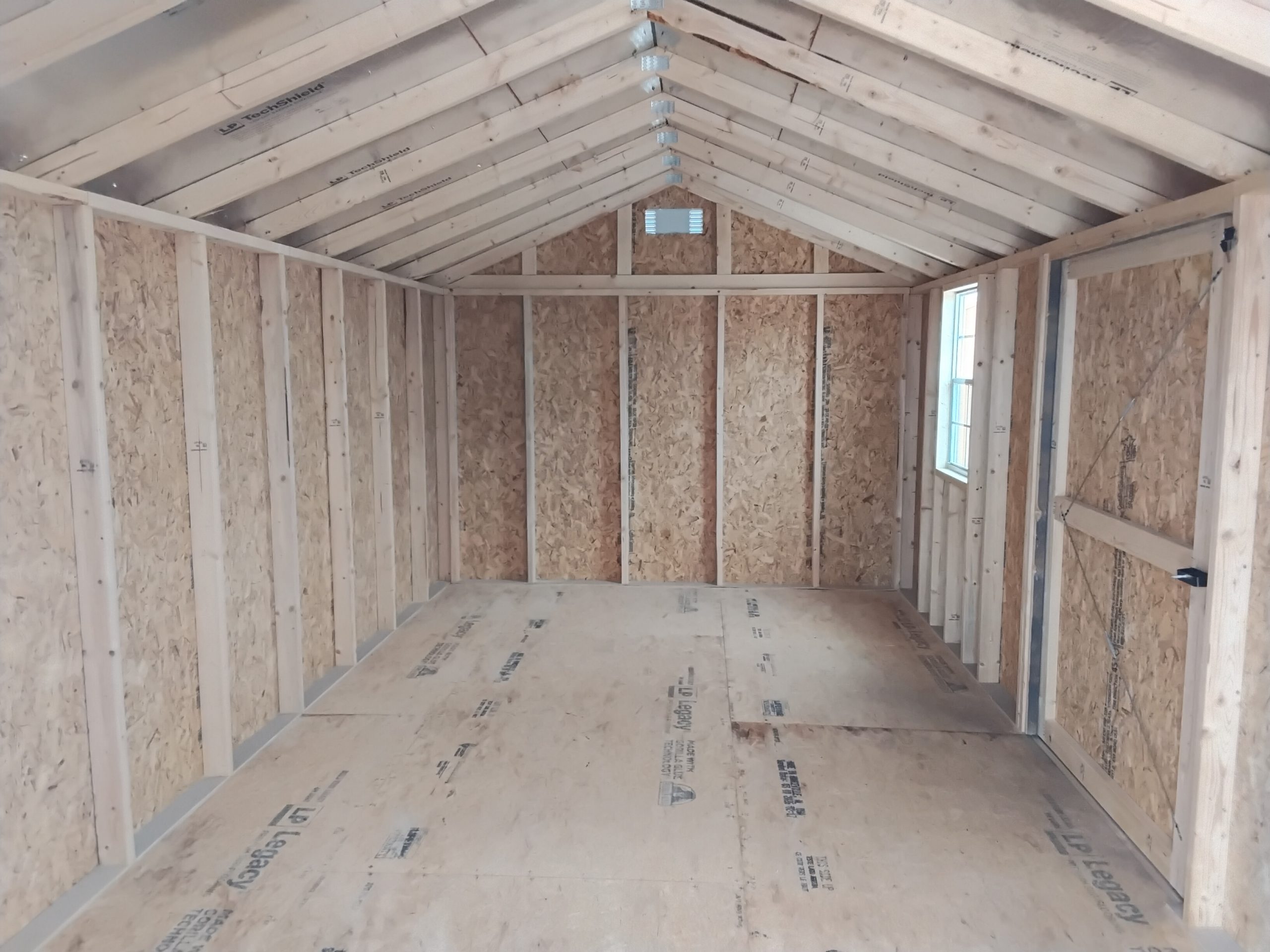 20260505_122808 - LELAND'S SHEDS - Unfinished interior room under construction with exposed wooden studs, OSB walls, and triangular roof trusses ending in a small window at the far wall.