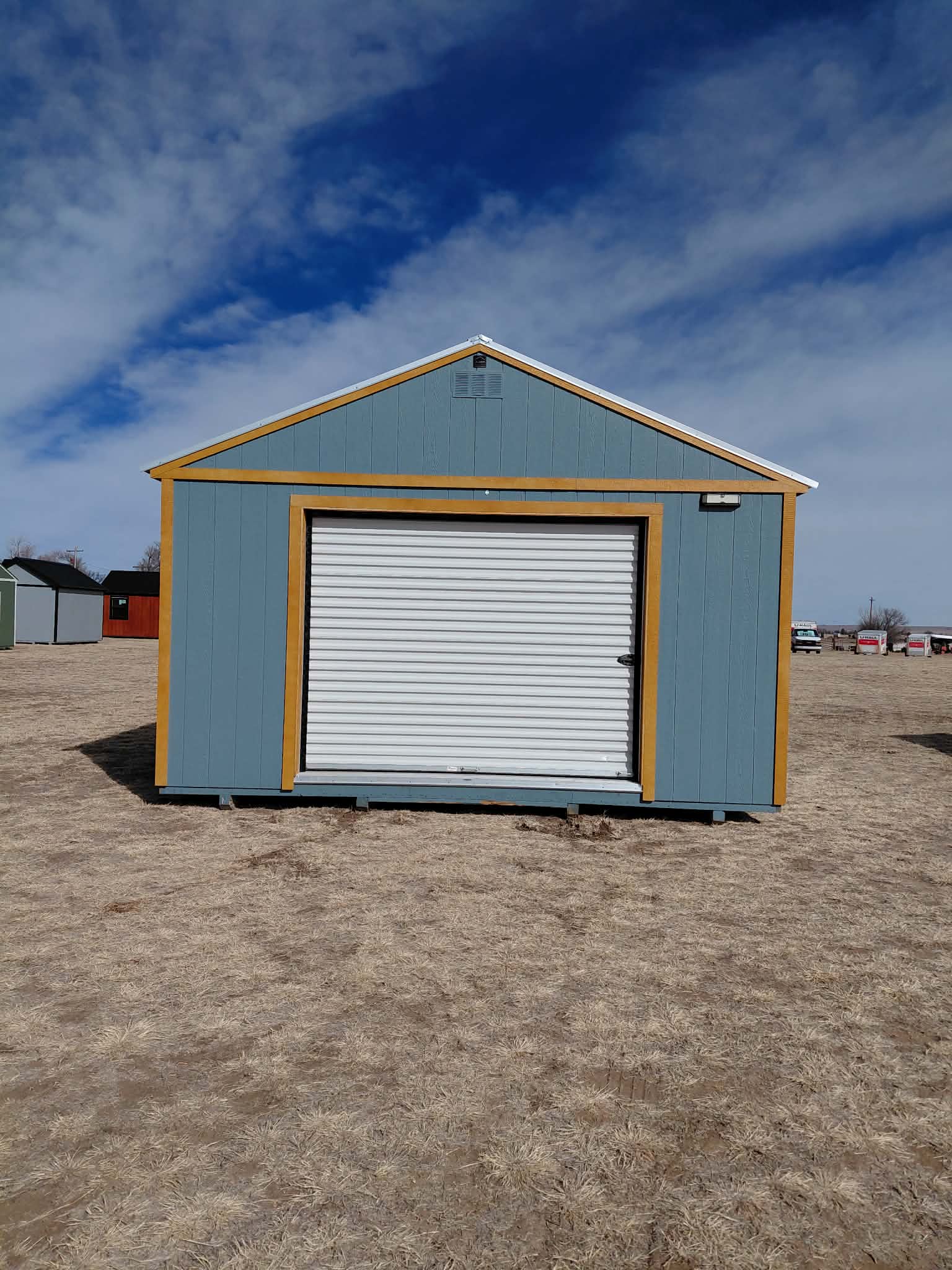  - Blue shed with yellow trim and a white roll-up door in a dry field, under a partly cloudy blue sky