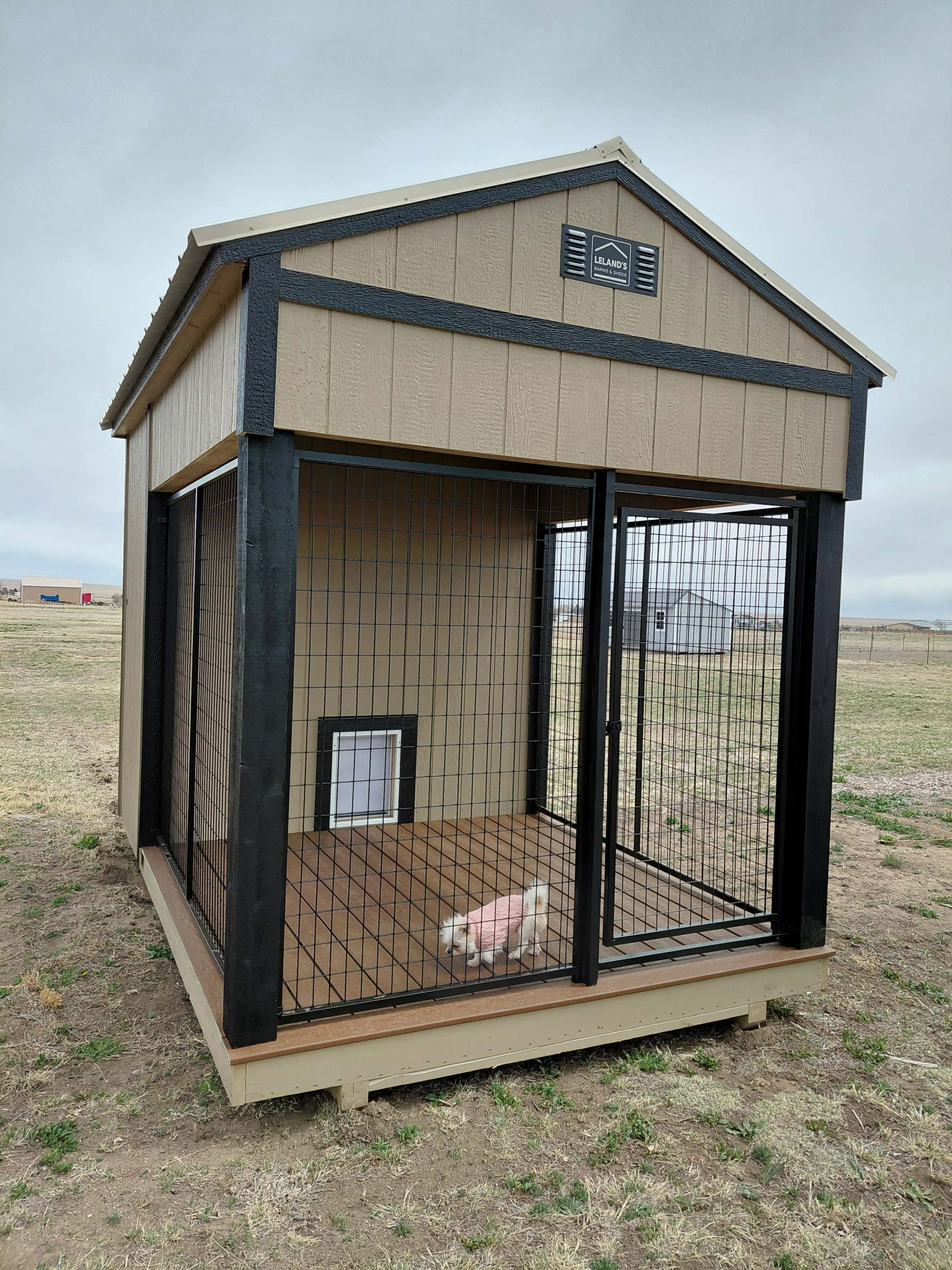  - A beige wooden dog kennel with black trim and wire fencing, housing a small white fluffy dog inside on a dirt yard.