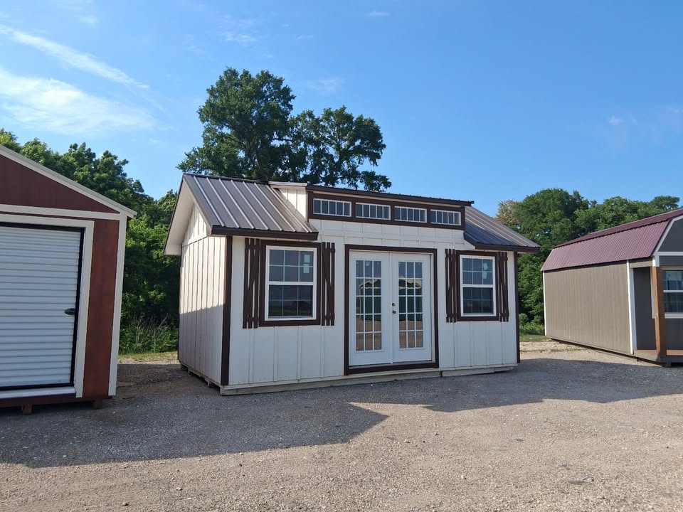  - A white shed with brown trim and French doors, two windows, and a metal roof in a sunny outdoor lot.