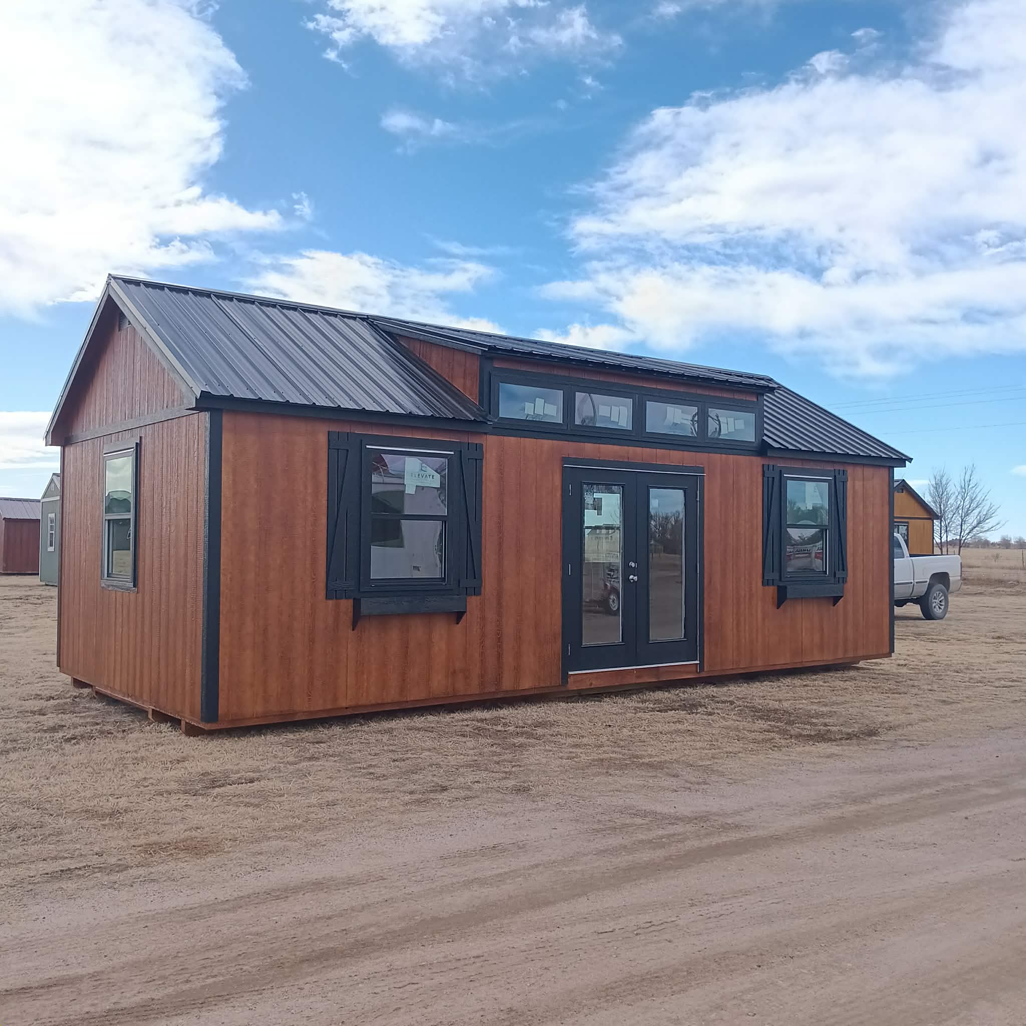  - Wooden one-story house with dark trim and a metal roof on a dirt lot beneath a blue sky with clouds in the background.