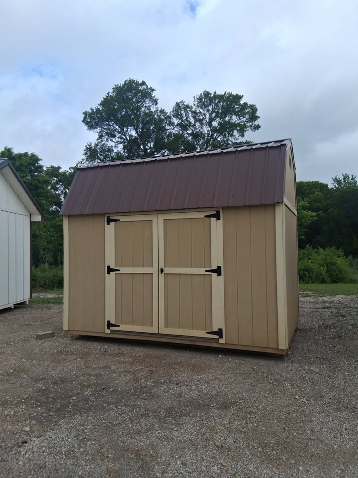  - Beige wooden storage shed with double doors and black hinges on a gravel lot, brown metal roof, trees behind.