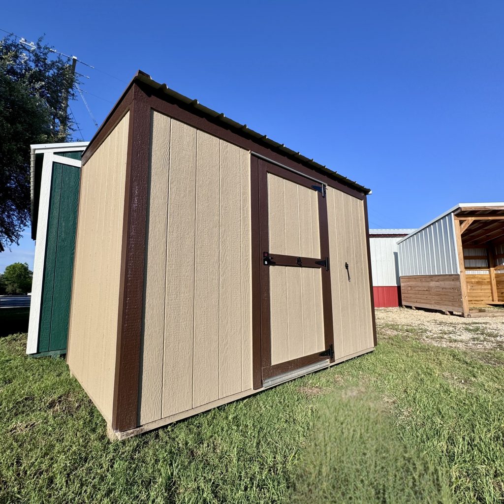  - Brown and tan wooden shed with green doors against a clear blue sky, standing on grass.