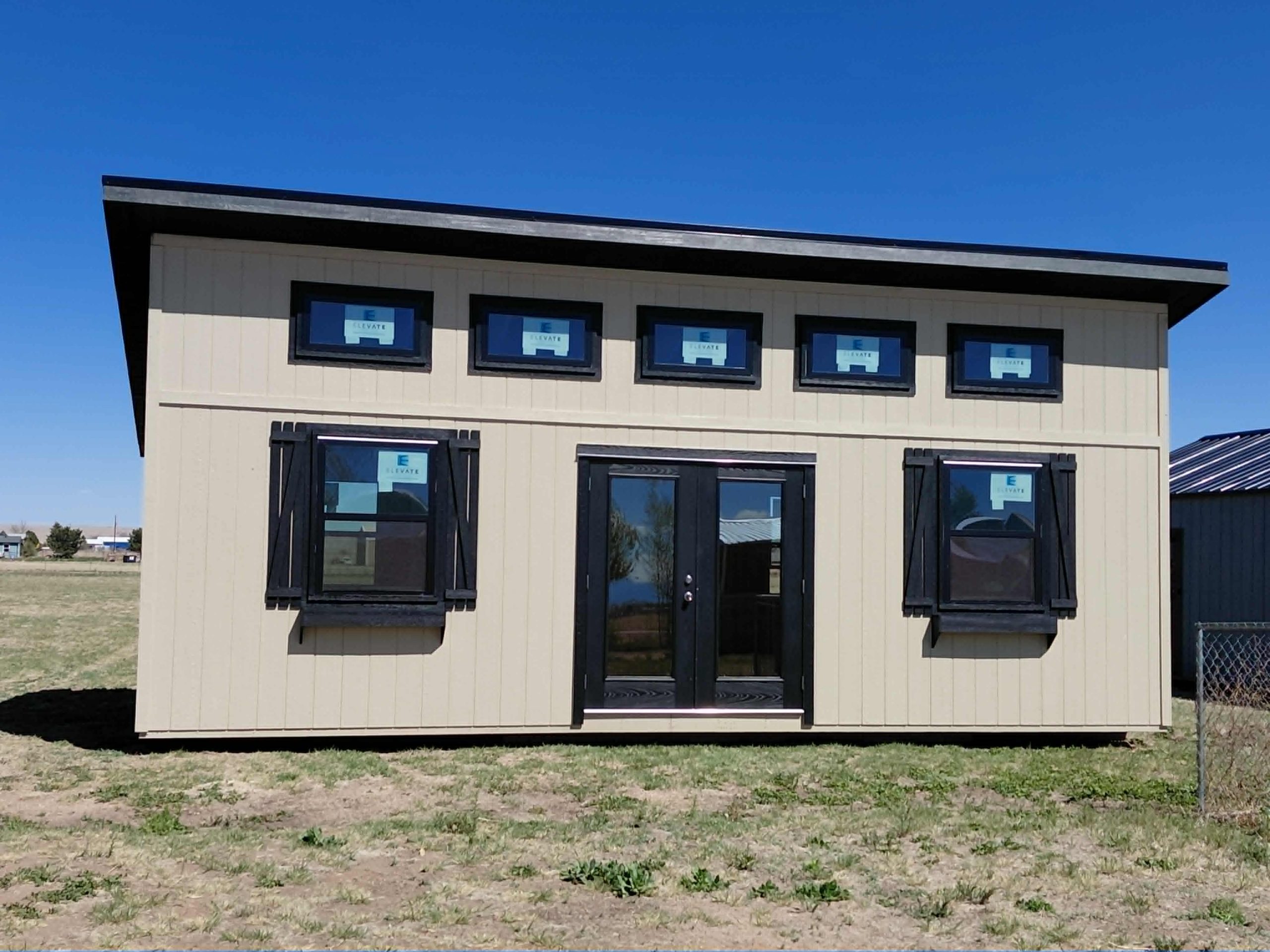  - Two-story beige building with large black-framed windows and a glass door under a flat roof, set on a grassy lot under a clear blue sky