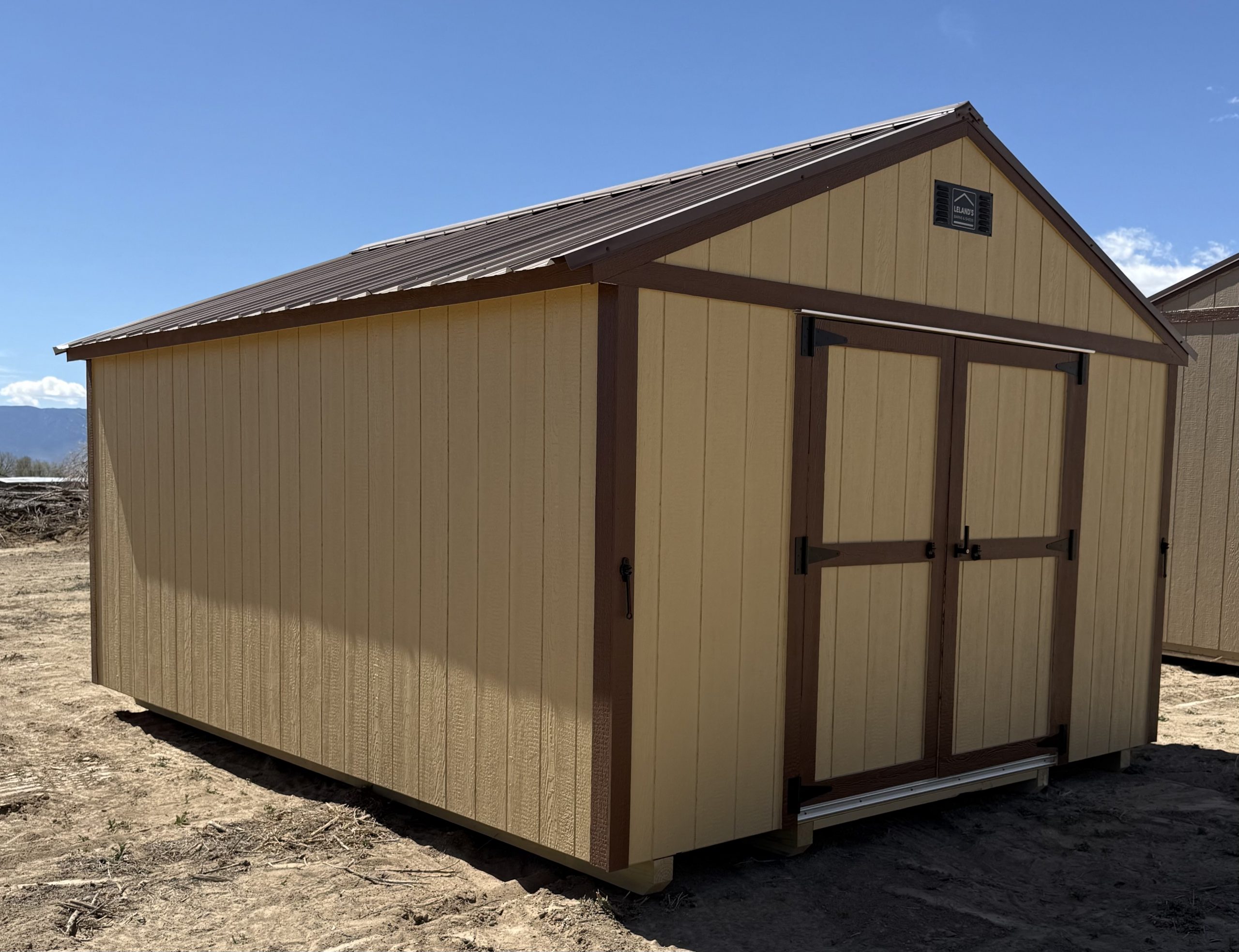  - Beige backyard storage shed with brown trim under a clear blue sky; vented gable and double doors on the side paneling.