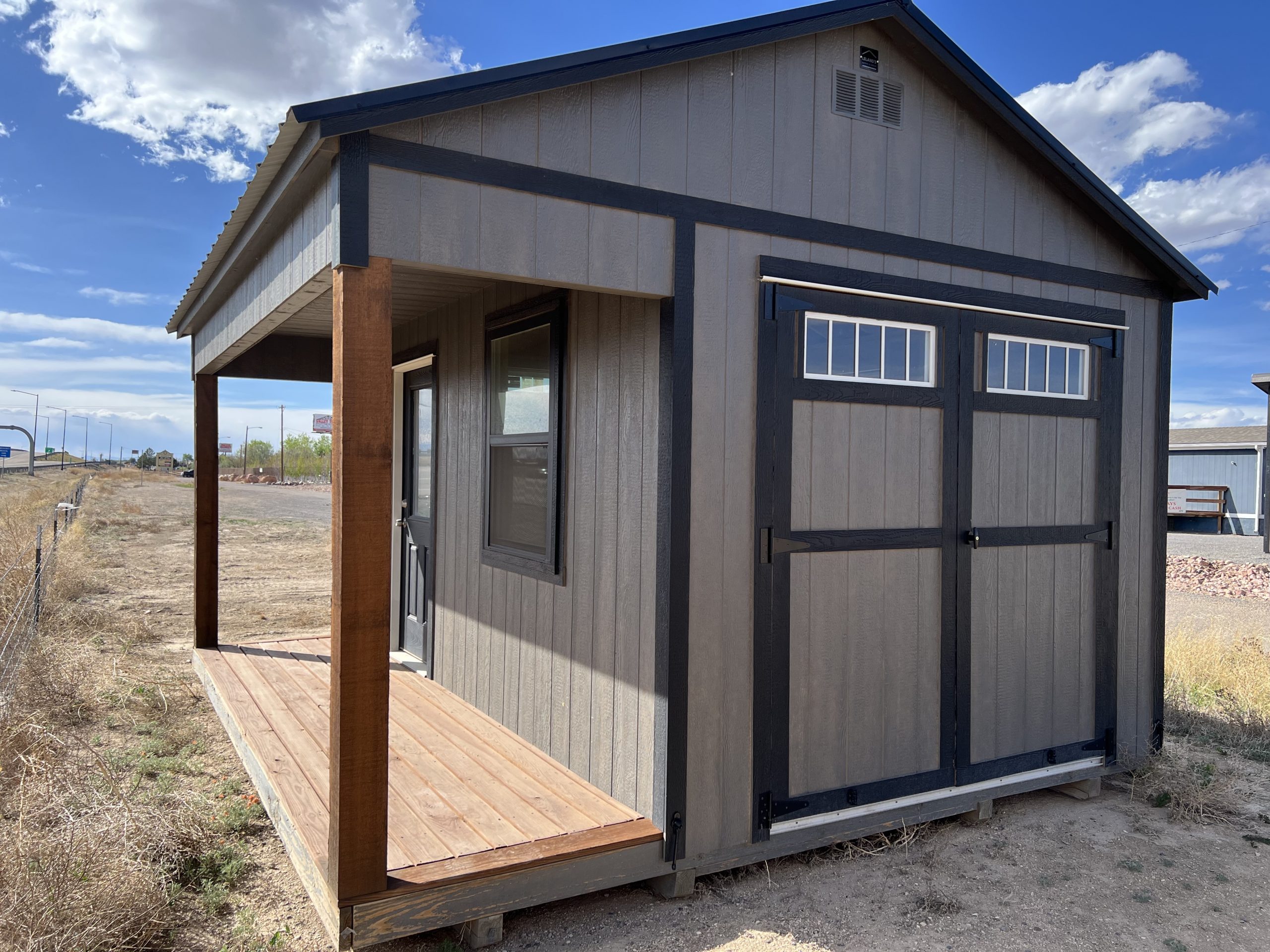  - Small gray shed with dark trim and a wooden front porch in a dry, open landscape under a blue sky.
