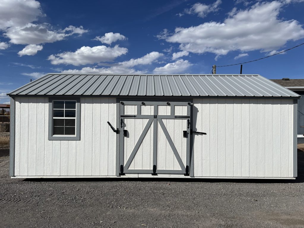  - White storage shed with a gray metal pitched roof, white exterior, and a large double-door with black hardware, set on gravel against a blue sky with clouds (left window visible).