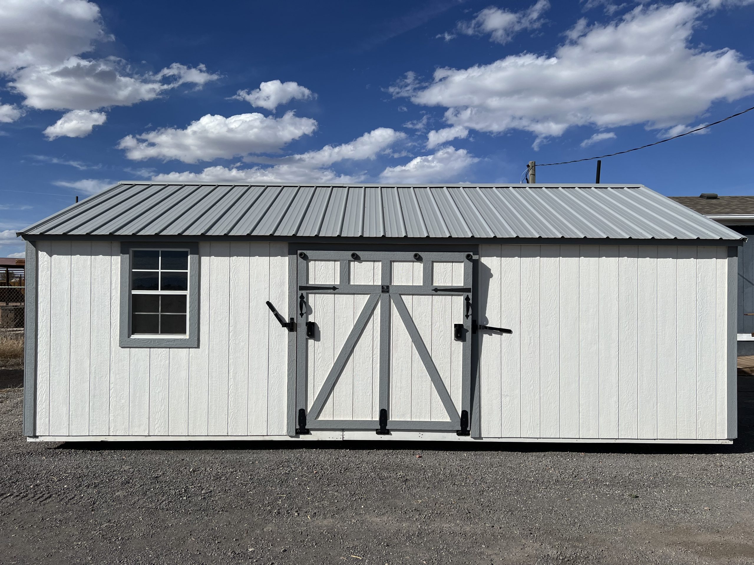 - White storage shed with a gray metal pitched roof, white exterior, and a large double-door with black hardware, set on gravel against a blue sky with clouds (left window visible).
