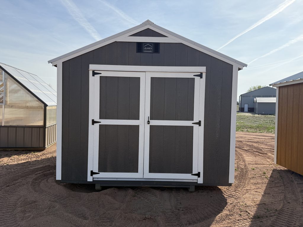  - Front view of a gray shed with white trim and double doors, set in a sunny yard with other outbuildings nearby.