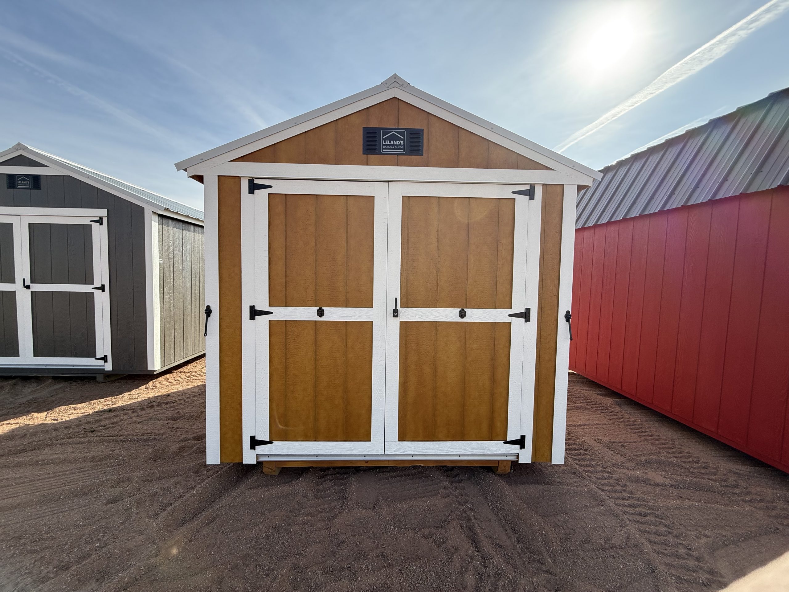  - A row of outdoor storage sheds with a central brown shed framed in white, set on dirt under a bright sun in a clear sky.