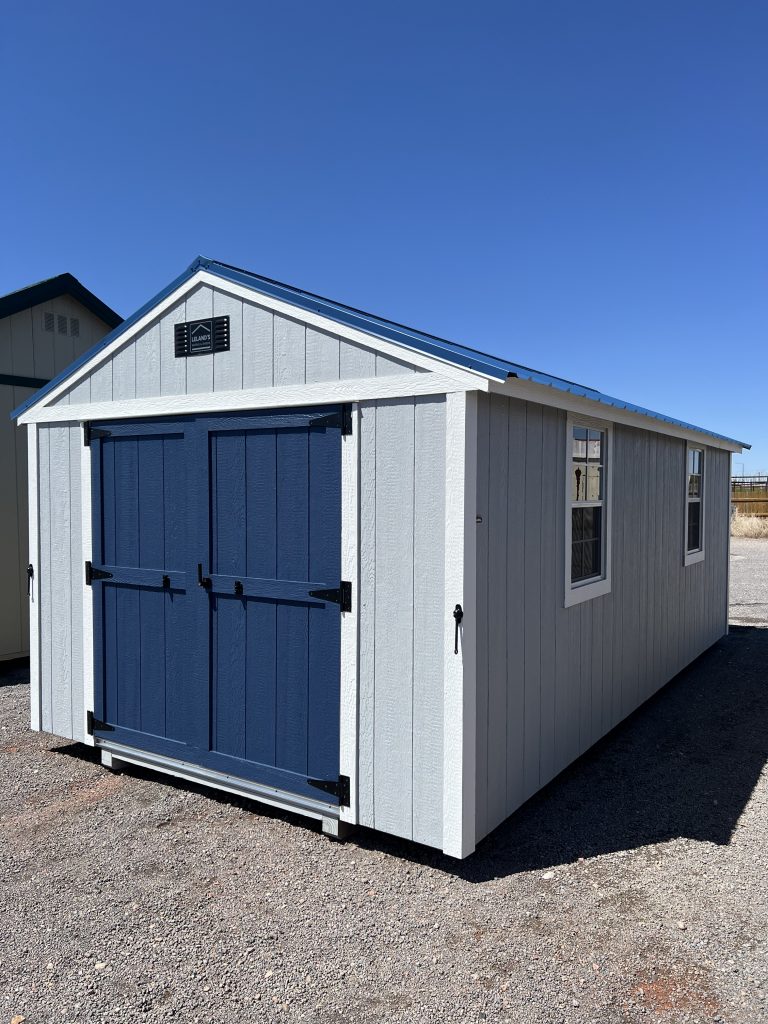  - White wooden shed with blue double doors and trim, set on a gravel lot under a bright blue sky.