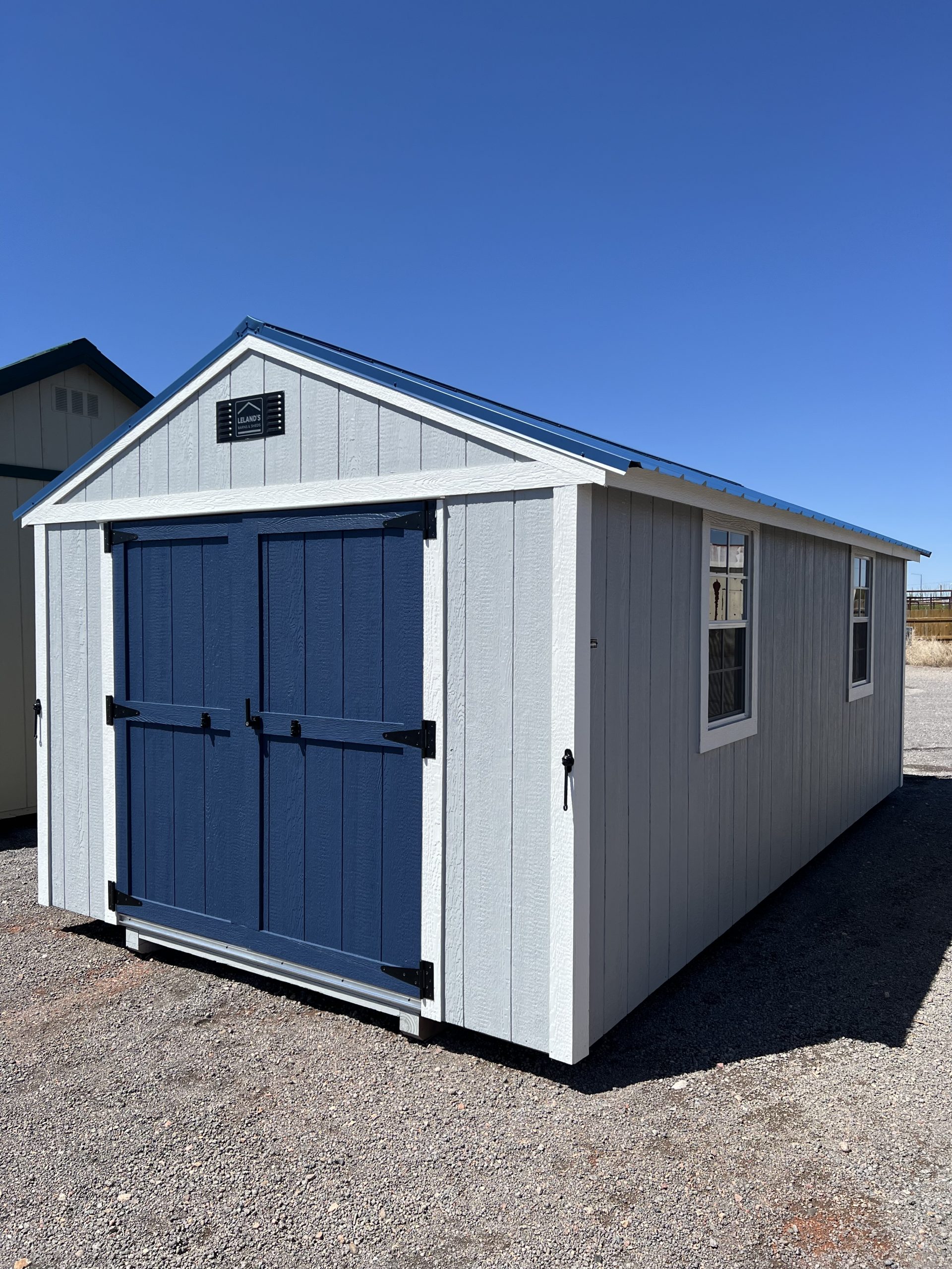  - White wooden shed with blue double doors and trim, set on a gravel lot under a bright blue sky.