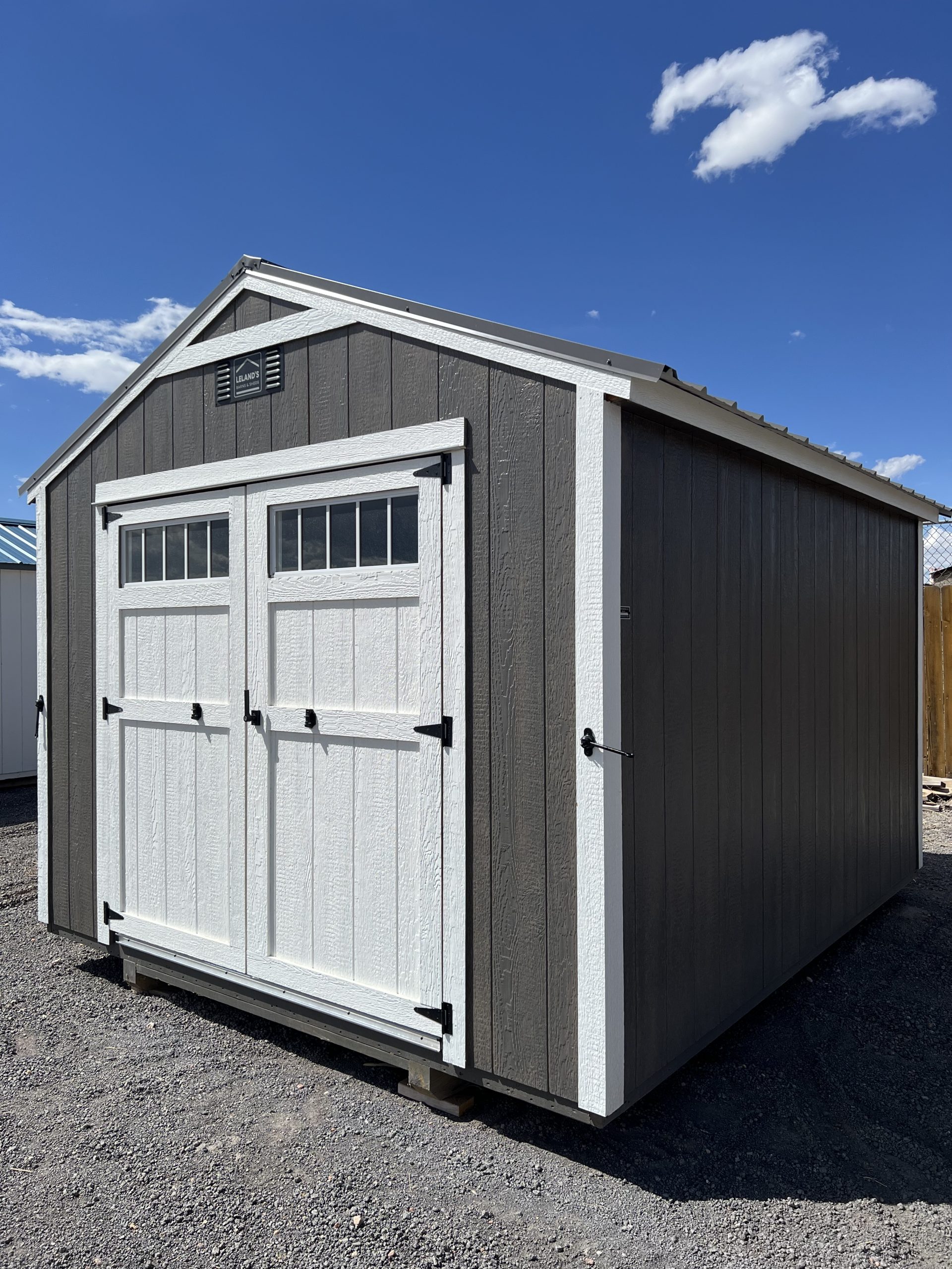 - - Grayscale shed with white-framed double doors on a gravel lot beneath a blue sky with a few clouds