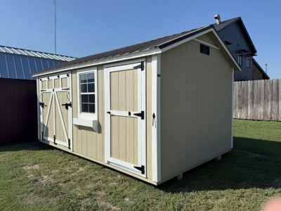IMG_2333 - LELAND'S SHEDS - Beige garden shed with white trim and black hardware in a grassy yard beside a wooden fence on a sunny day.