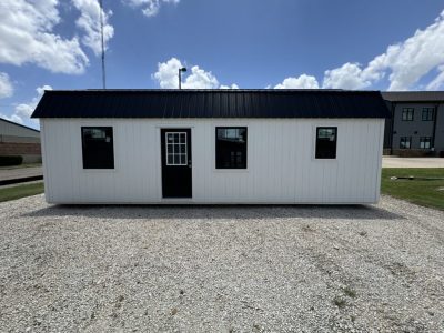 IMG_3855 - LELAND'S SHEDS - White prefabricated shed with a black metal roof on a gravel lot, featuring three windows and a front door with a divided-glass panel.