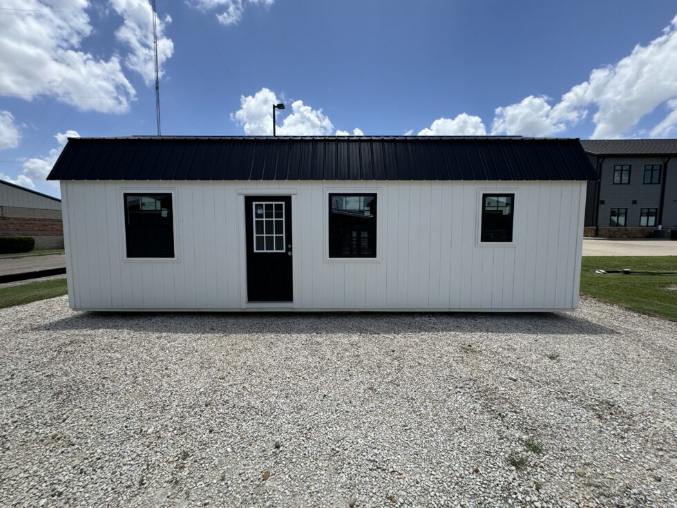 - - White prefabricated shed with a black metal roof on a gravel lot, featuring three windows and a front door with a divided-glass panel.