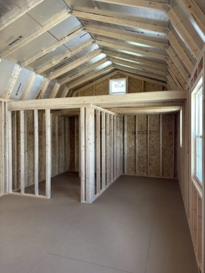 IMG_3861 - LELAND'S SHEDS - Interior of a partially built home with exposed wooden studs, framed walls, and a pitched roof with foil insulation.