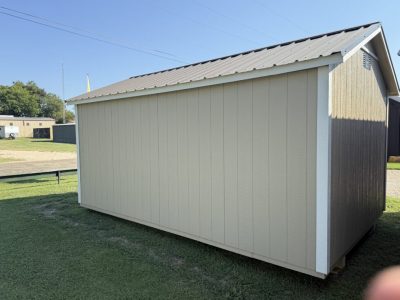 IMG_3864 - LELAND'S SHEDS - Side view of a beige shed with white trim and metal roof on a grassy lot, clear blue sky above.